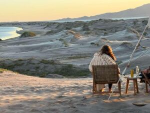 Photo of a woman sitting in a chair on a sand dune next to her glamping tent overlooking Magdalena Bay in Baja, Mexico.